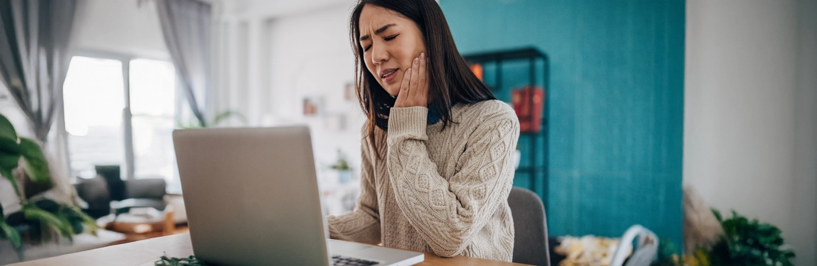 A woman who has a toothache, holding her jaw with a pained expression while looking at a laptop screen